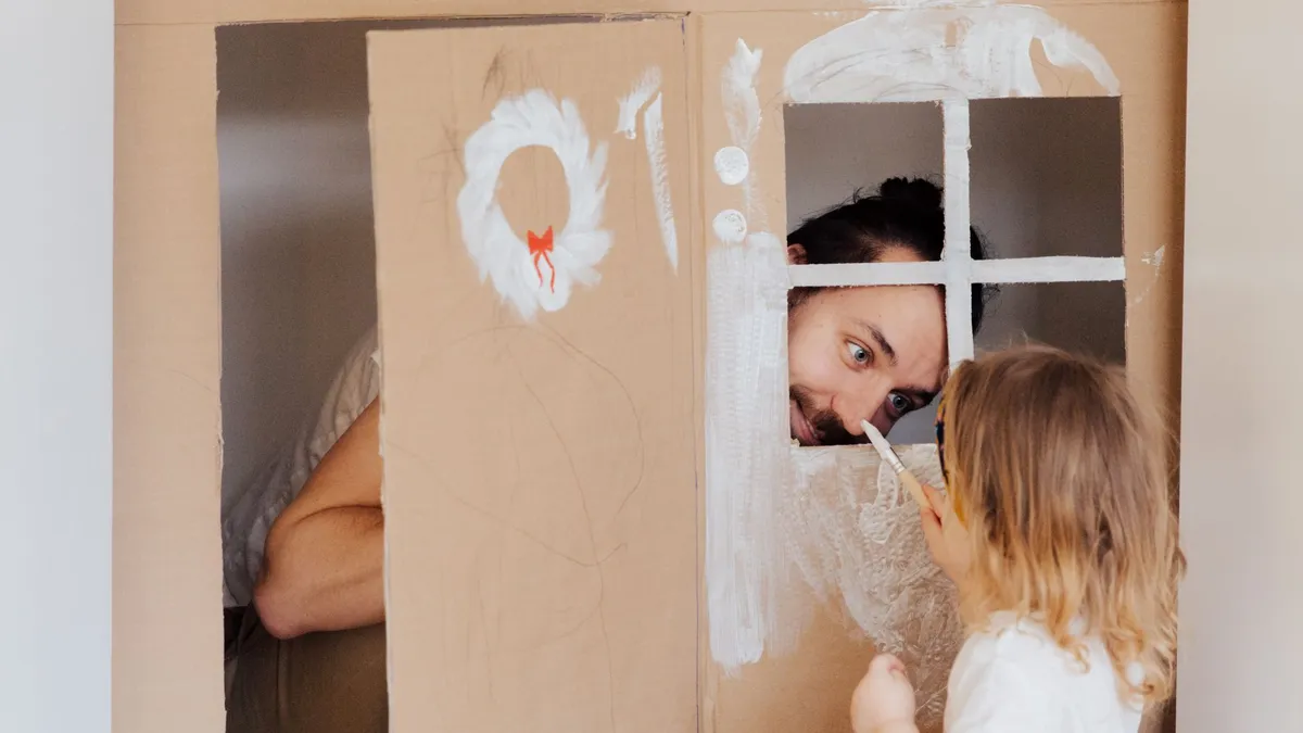 Un parent et sa fille peignent ensemble une maison en carton dans le salon